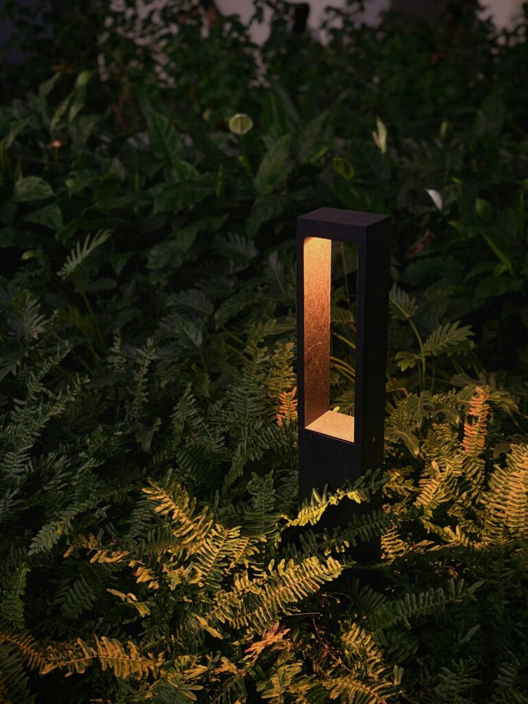 Moderne Gartenleuchte wirft warmes Licht auf grünes Laub – insektenfreundliche Gartenbeleuchtung