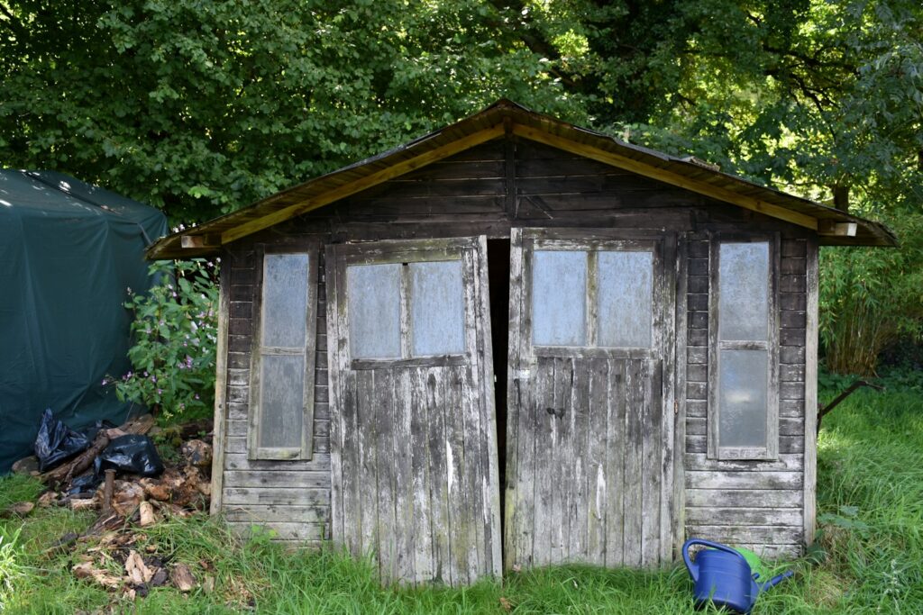 Verwittertes Gartenhaus aus Holz mit Glasfenstern, das dringend Pflege braucht