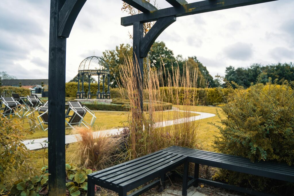 Blick durch eine dunkle Holzpergola mit Bank auf Ziergräser und einen Gartenweg – Markise oder Pergola als Schattenlösung im Garten